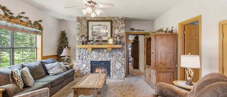 A cozy living room with a stone fireplace, a ceiling fan, and a large window.