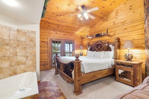 A wooden cabin bedroom with a bed, a nightstand, a lamp, a ceiling fan, and a stone wall.