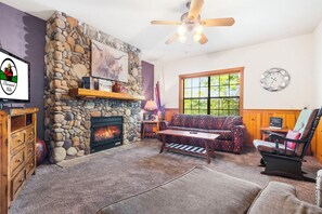 A cozy living room with a stone fireplace, a wooden ceiling fan, a patterned sofa, and a window with a view of greenery.