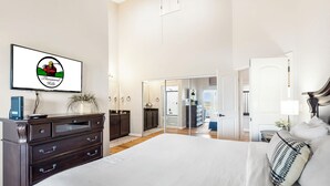 Un dormitorio amplio con una cama grande, un aparador, un televisor mostrando un logo y un espejo que refleja la habitación.