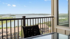 Un balcón con vistas a un río y colinas lejanas, una mesa de vidrio y una silla negra.