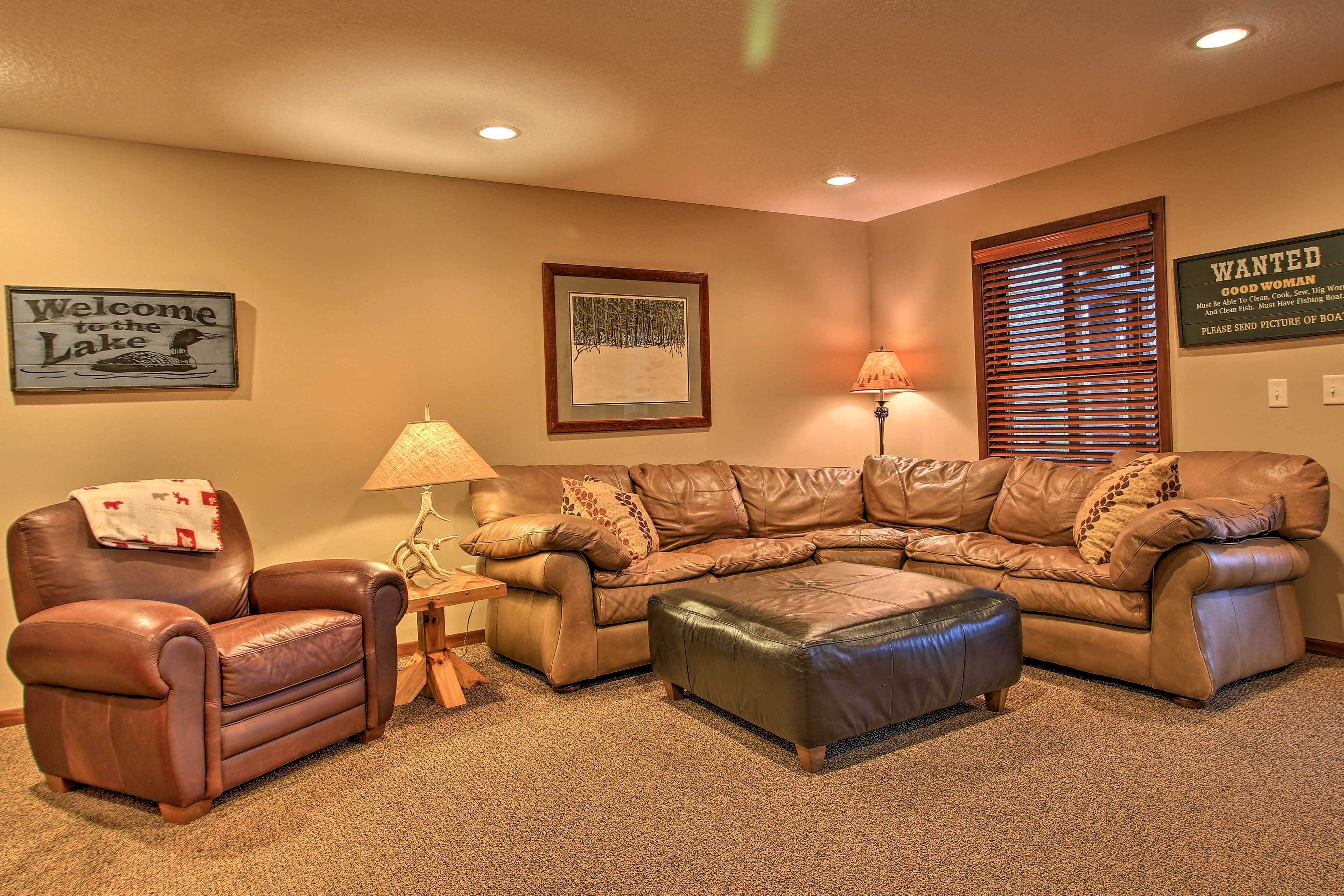 A living room with a brown leather sofa, a leather armchair, and a matching ottoman. There is a lamp on a side table, a framed picture on the wall, and a 'Welcome to the Lake' sign.