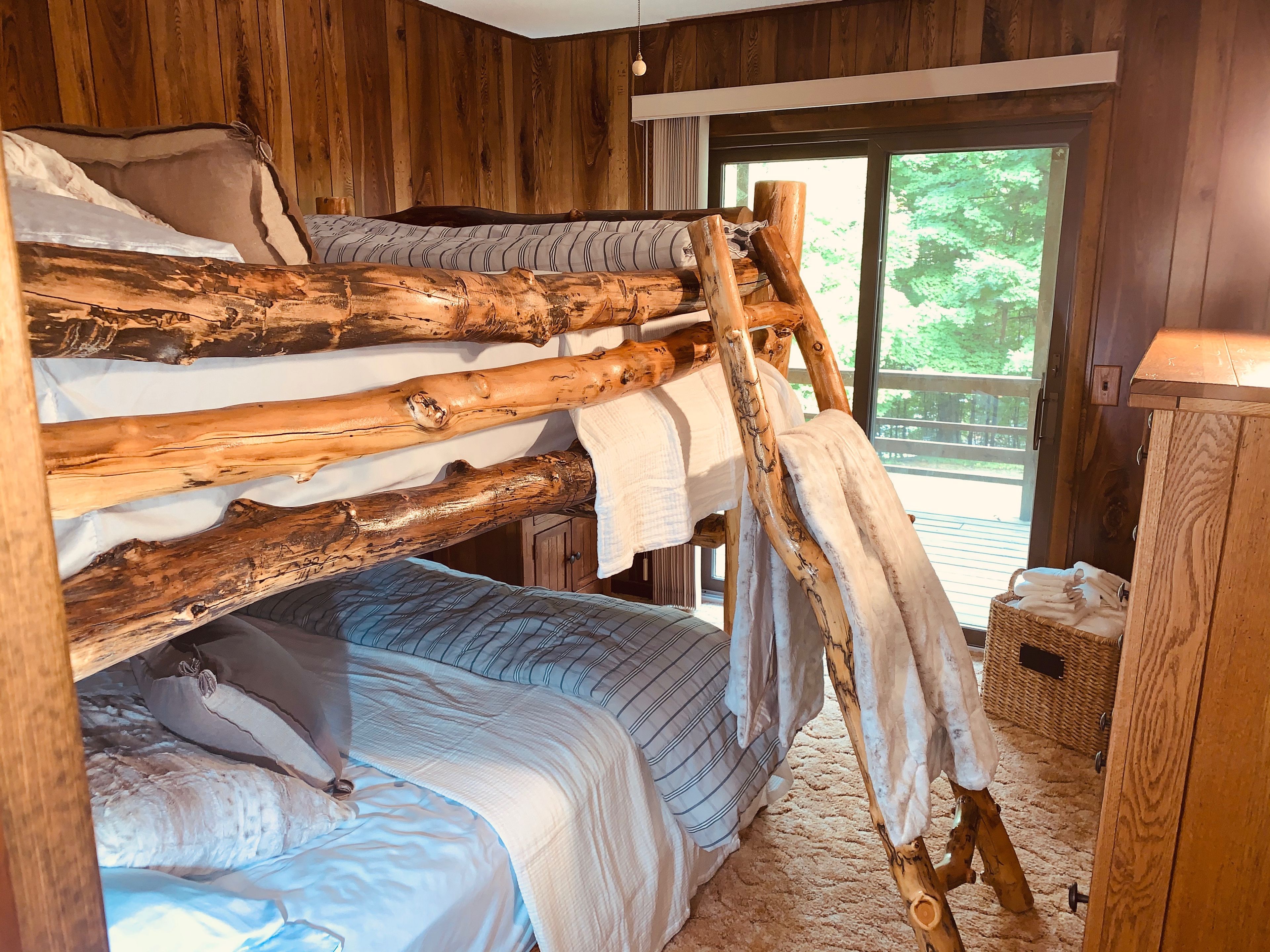 Una cama litera con una escalera de madera, una ventana que da a la vegetación y una cesta de mimbre con toallas.