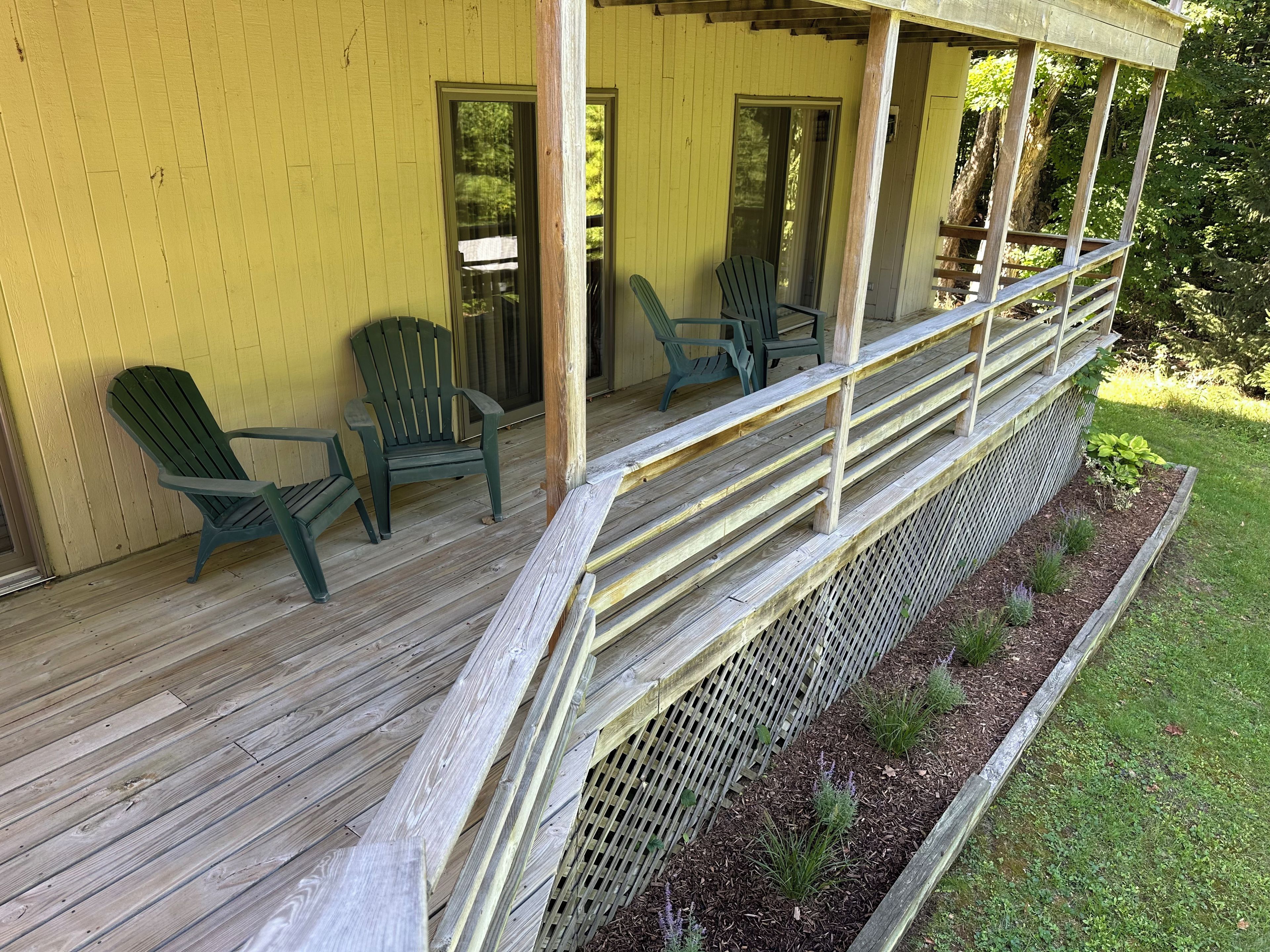 Una terraza de madera con sillas Adirondack y una pequeña mesa, rodeada por una cerca de madera y un cantero con plantas.
