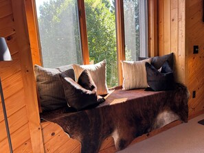 A wooden bench with cushions against a window, overlooking a forest.