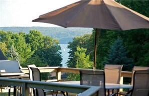 Une terrasse extérieure avec une table et des chaises, un grand parasol et une vue sur un lac entouré d’arbres.