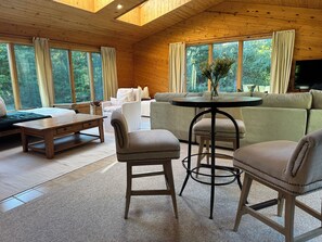Un salón acogedor con sofá, dos sillones, una mesa de centro y televisor. Amplias ventanas ofrecen vistas al exterior.