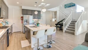 A modern kitchen with a central island, white bar stools, and a piano in the living area. - Lake & Mountain Views: Indio Oasis w/ Pool (Indio)