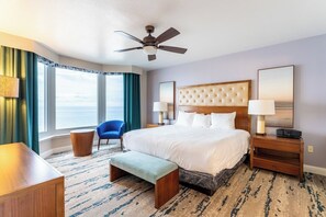 Une chambre d’hôtel avec un grand lit, un ventilateur de plafond, une vue sur l’océan, un fauteuil bleu et une table de chevet en bois avec une lampe.