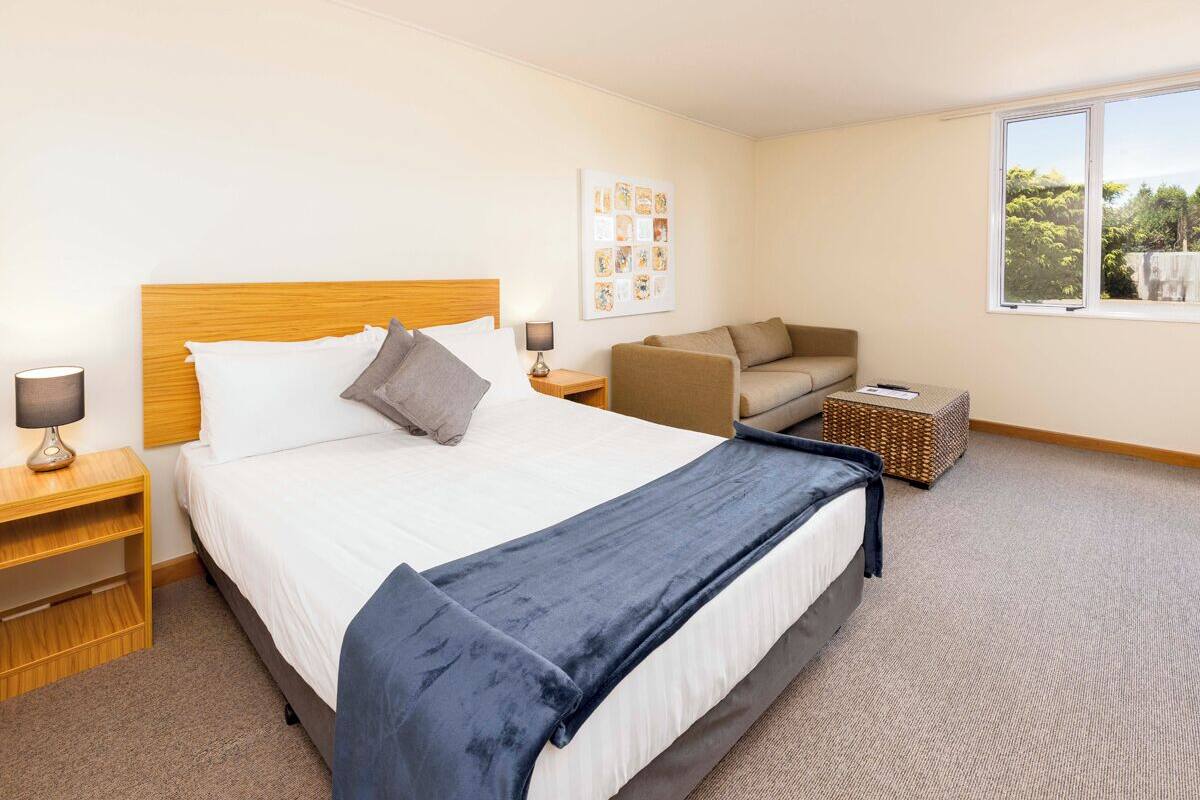 A hotel room with a large bed, a sofa, a small table, a lamp, and a window with a view of greenery.
