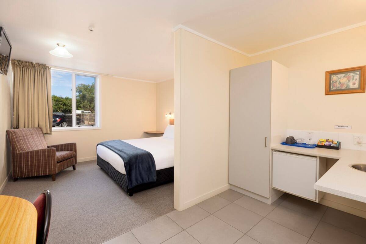 A compact hotel room with a bed, a small desk, a chair, a window with curtains, and a kitchenette.