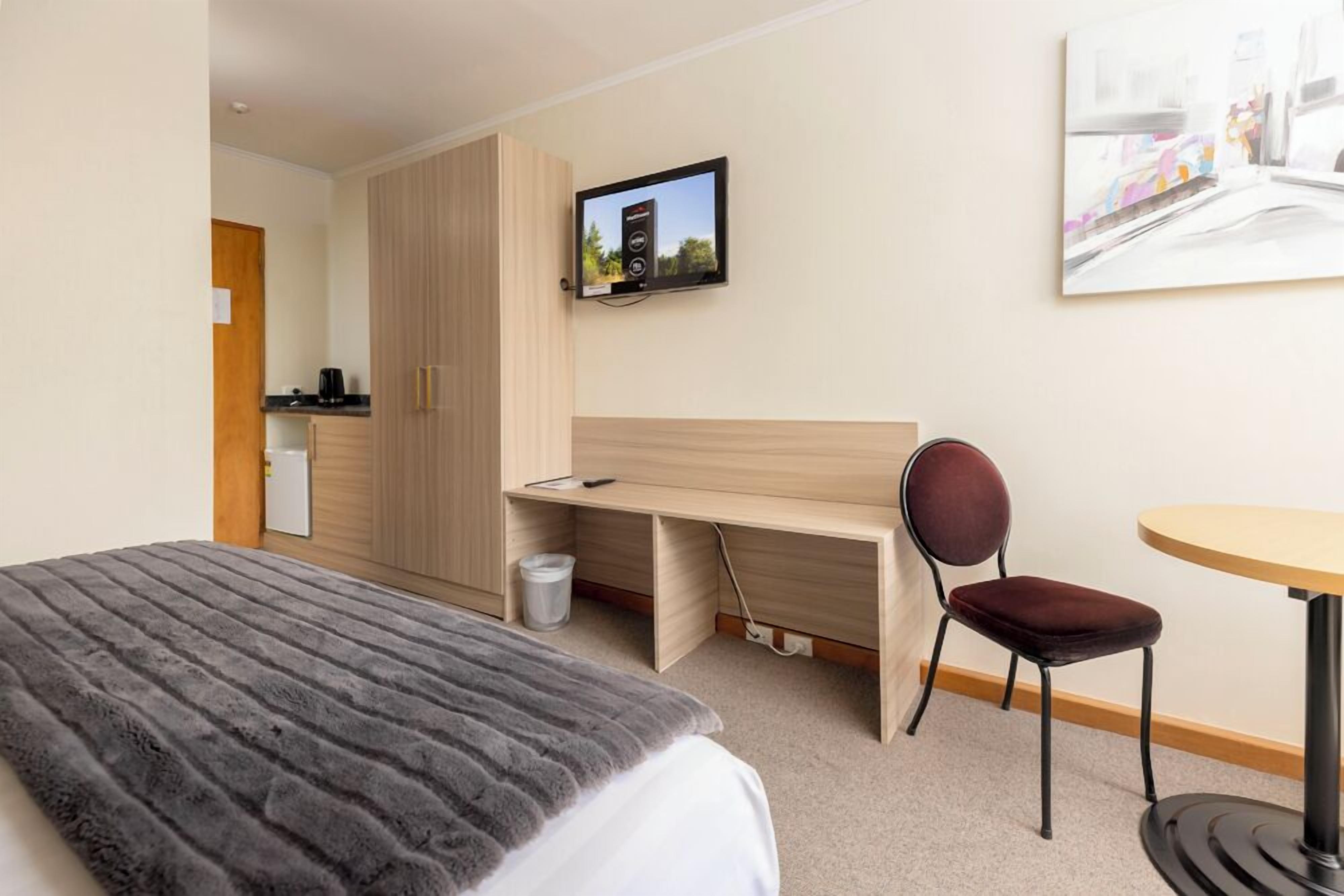 A hotel room with a bed, a desk, a chair, a TV, and a painting on the wall.