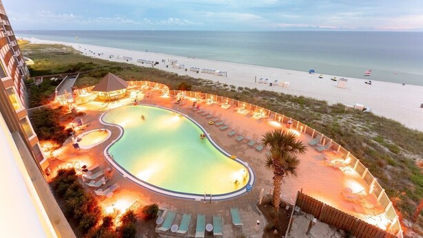 Ein Pool mit geschwungener Form, umgeben von Liegestühlen und einem Holzzaun, mit Strand und Meer im Hintergrund.
