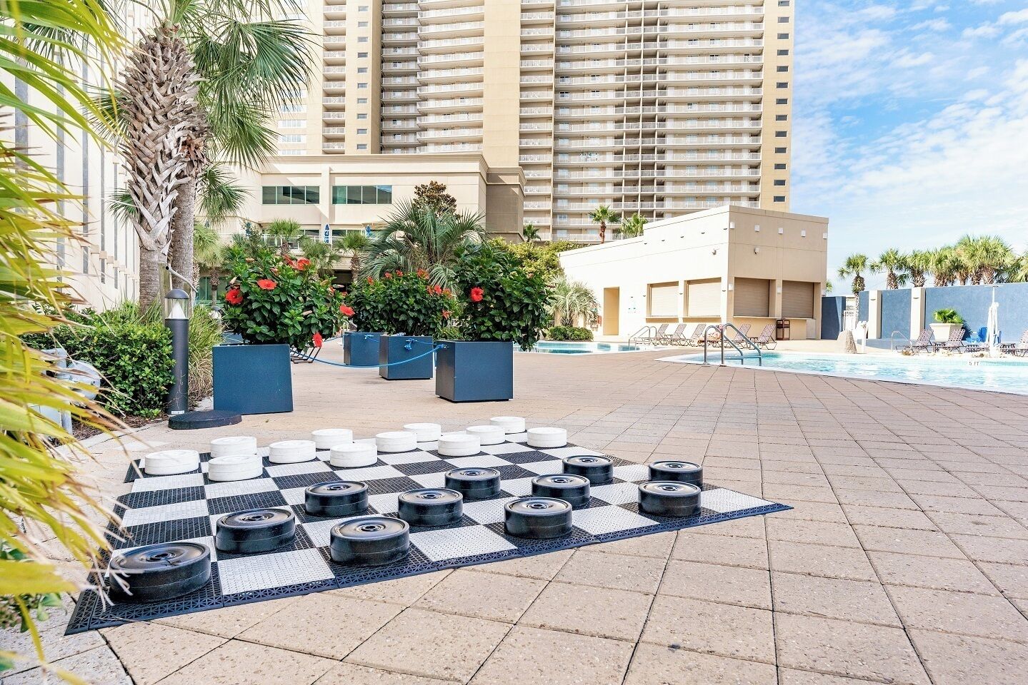 Un área de piscina con un diseño a cuadros hecho con neumáticos y plantas en macetas, una zona de descanso junto a la piscina, y un edificio alto al fondo.
