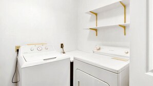 A laundry room with two white washers and dryers, a gold-colored shelf unit, and a white door. - Carole Cottage - close to golf, tennis, and the beach (Jekyll Island)