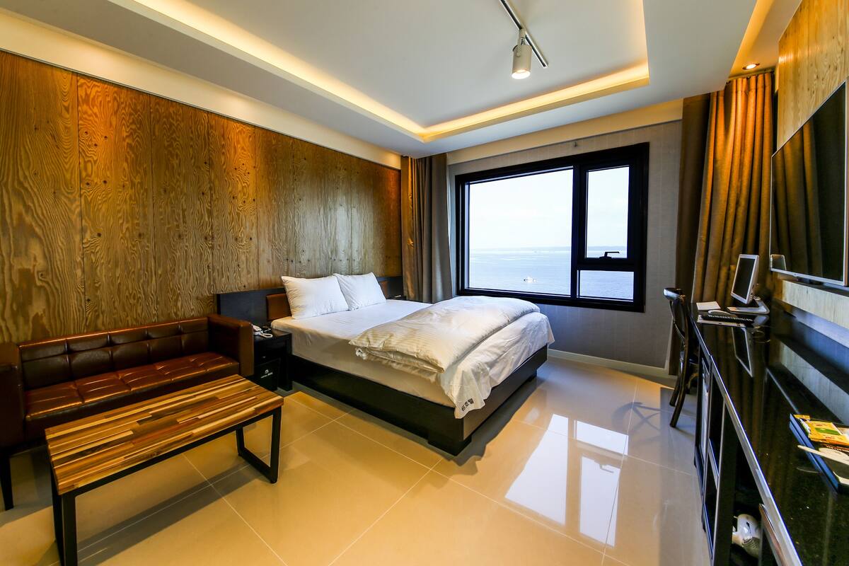 A modern hotel room with a large bed, a wooden bench, a desk with a computer, and a view of the sea.