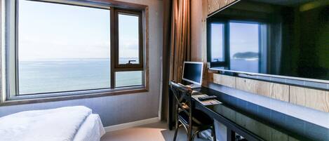 A hotel room with a large window, a bed, a desk with a computer, and a flat-screen TV.