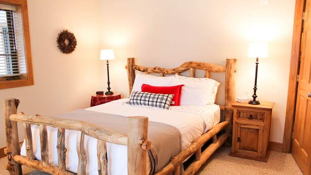 A rustic wooden bed with a headboard, two bedside tables with lamps, a small nightstand, and a wall-mounted wreath. - Boyne Mountain, Disciples Village Condo - Steps To Boyneland Run (Boyne falls)