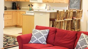 A red sofa with patterned cushions, a matching ottoman, a kitchen area with wooden cabinets and a microwave, and a chalkboard sign. - Boyne Mountain, Disciples Village Condo - Steps To Boyneland Run (Boyne falls)