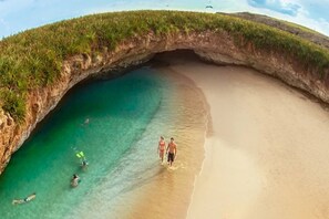 Vista aérea de una playa con una formación rocosa en arco, agua turquesa cristalina y una orilla arenosa.