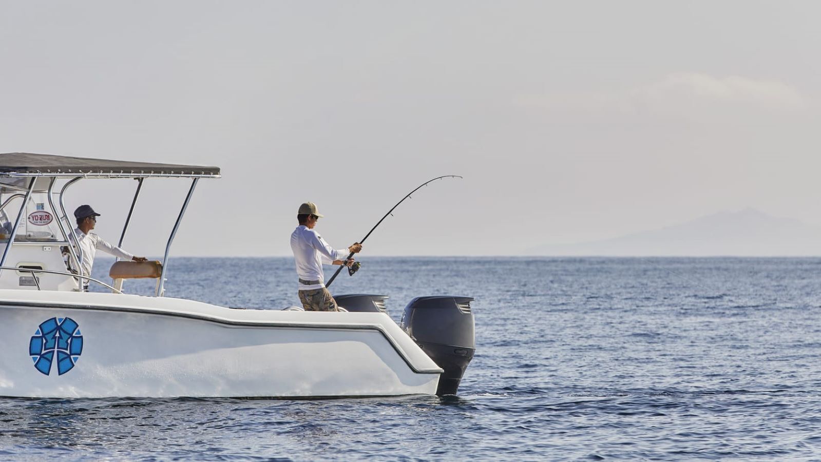Un bote con dos personas pescando, un logotipo en el costado y agua tranquila.