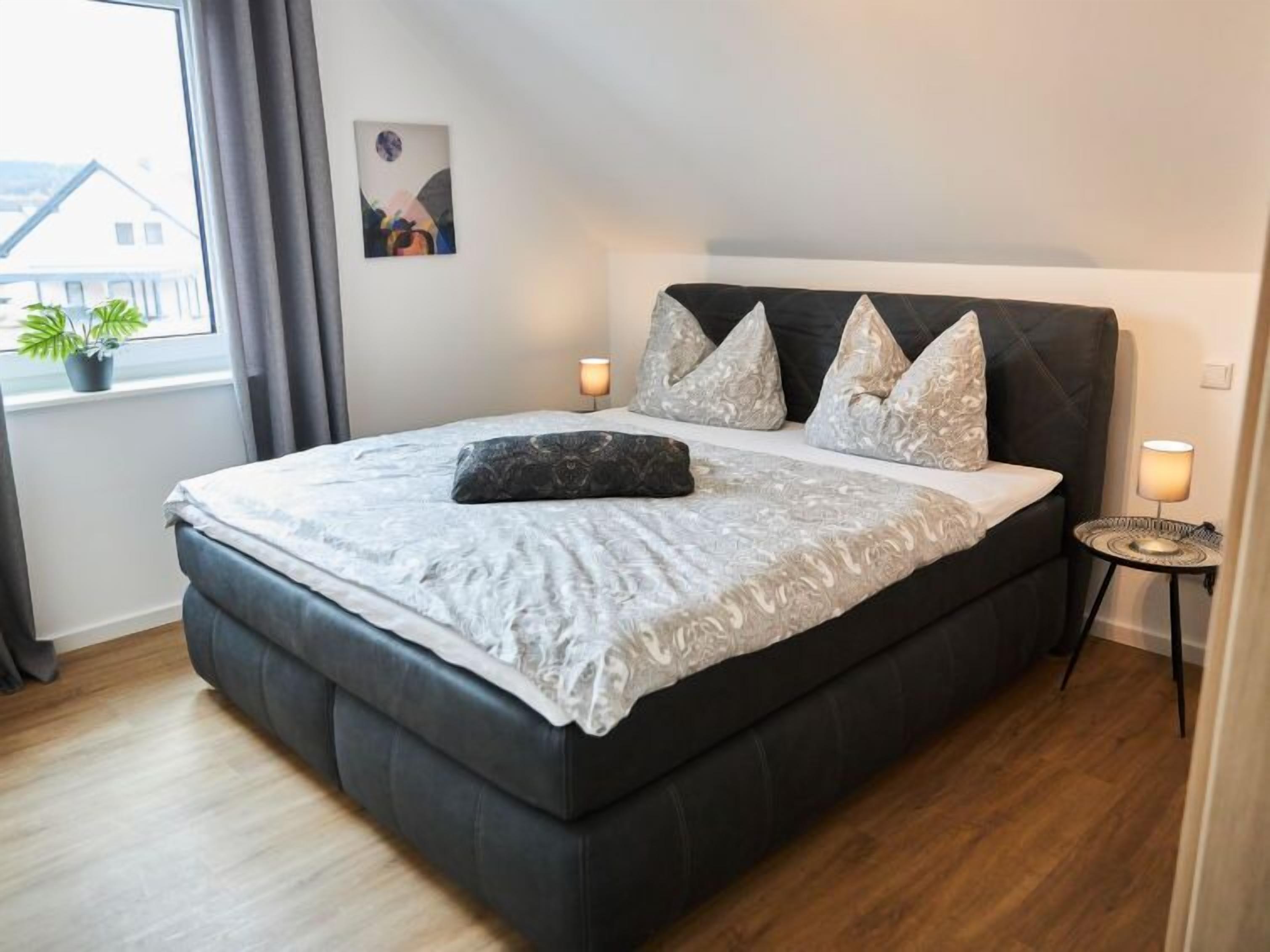 Una camera da letto moderna con un letto grande, biancheria grigia e cuscini decorativi. Si nota un comodino con una lampada, una pianta in vaso vicino alla finestra e un quadro appeso al muro.