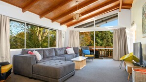 Un salon moderne avec un canapé d’angle, une table basse et un téléviseur. De grandes fenêtres et un balcon offrent une vue sur les arbres.