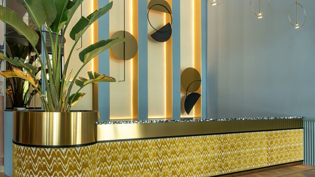 Un área de recepción con diseño moderno, que incluye un mostrador de recepción, una planta grande en maceta y una pared con motivos decorativos.