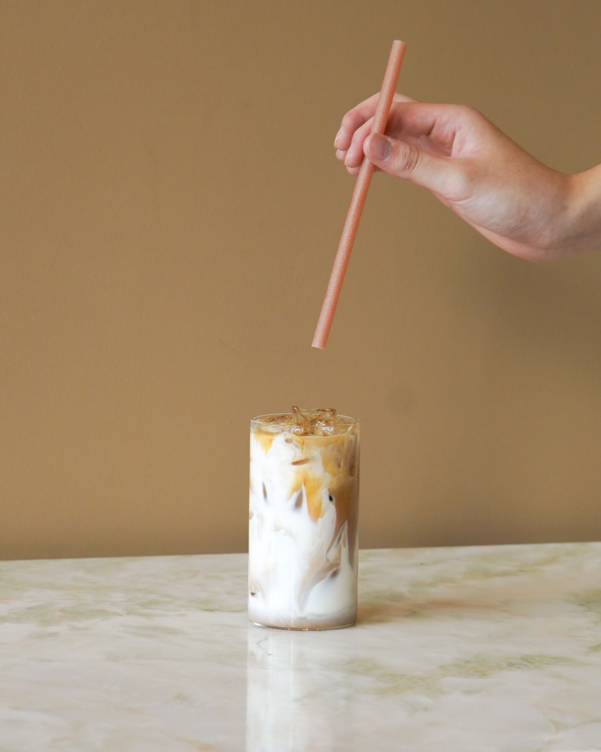 A hand holding a straw above a glass of iced coffee with a creamy layer and ice cubes.