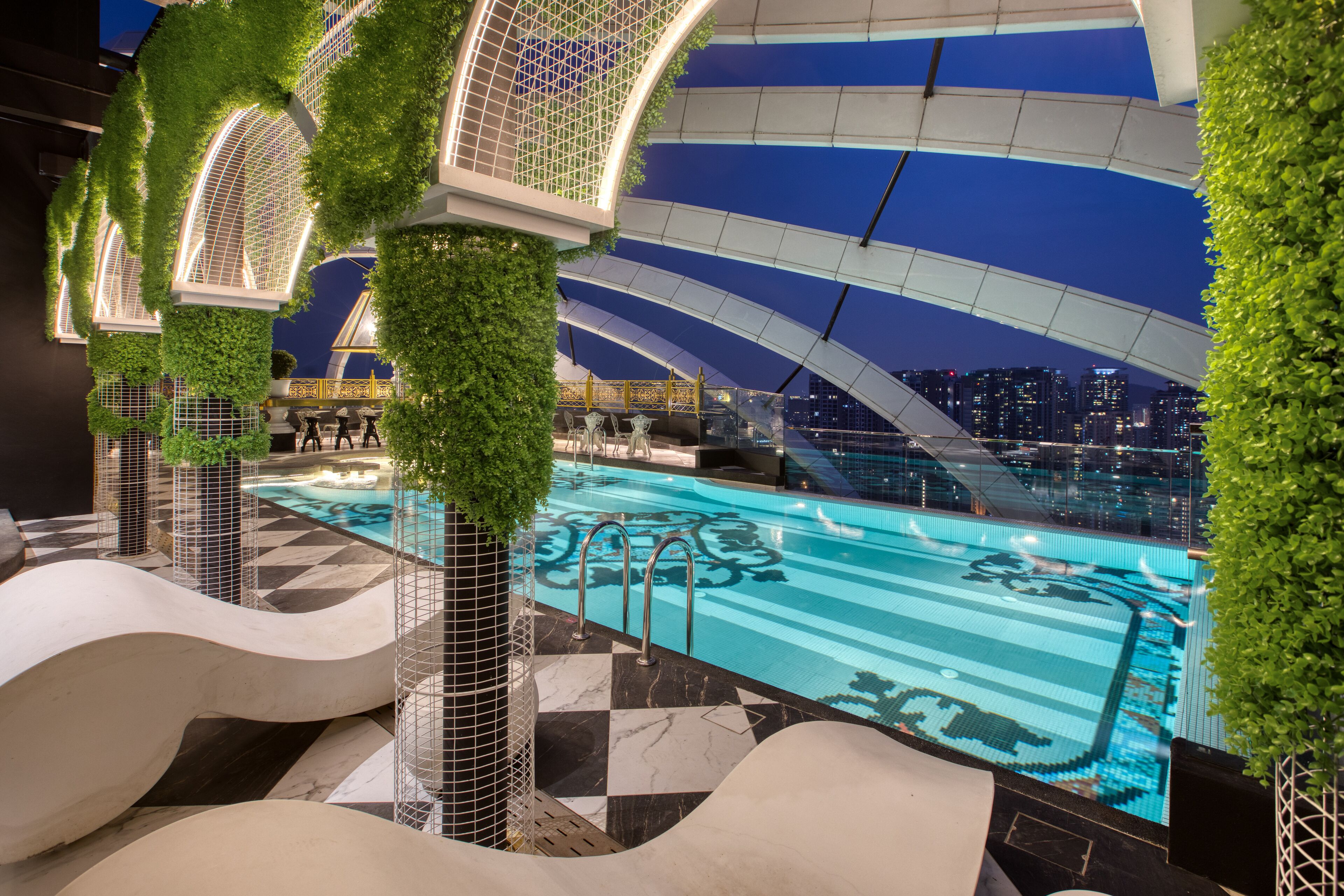 A modern indoor swimming pool with a cityscape view, surrounded by greenery and unique architectural elements.
