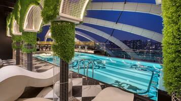 A modern indoor swimming pool with a cityscape view, surrounded by greenery and unique architectural elements.