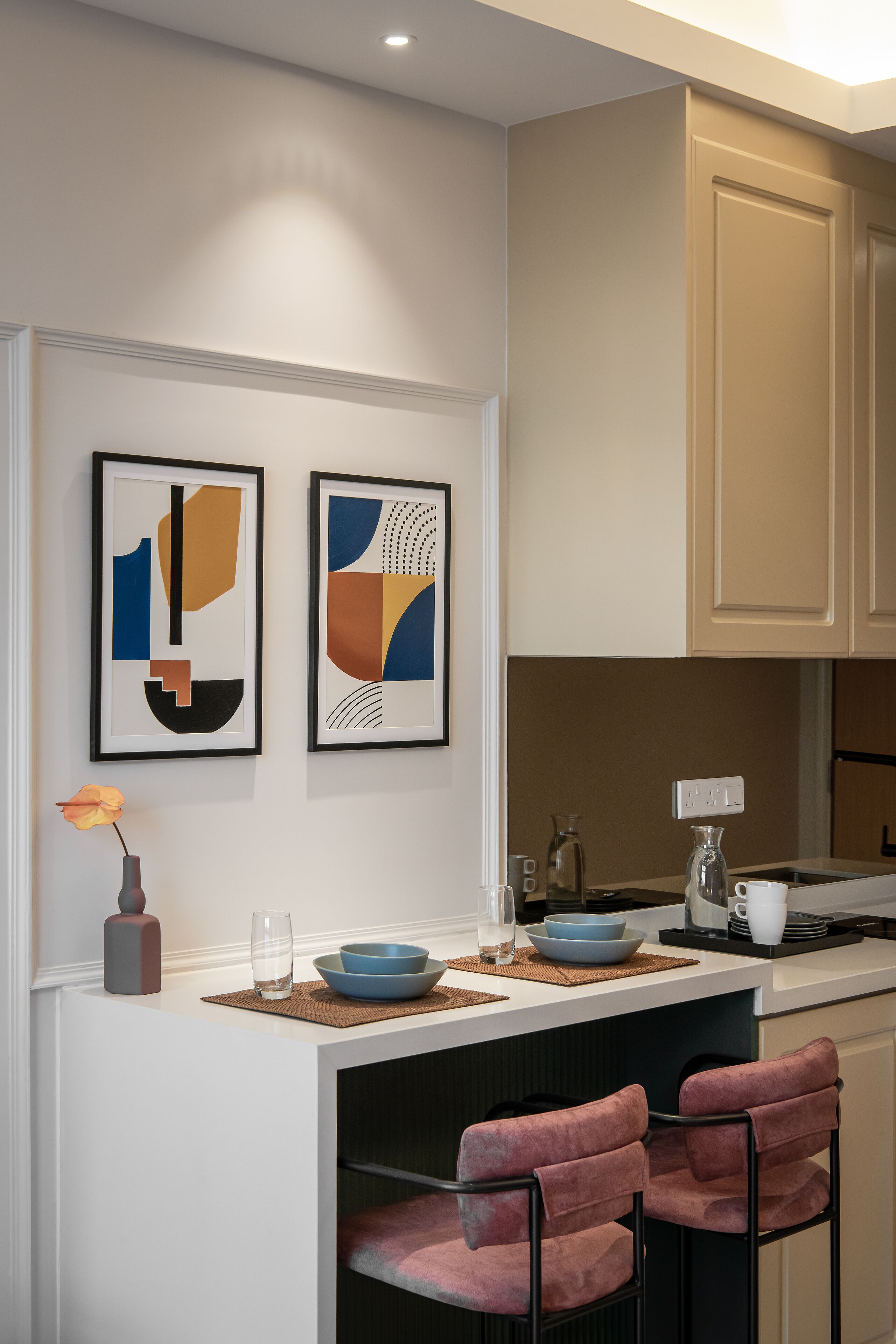 A modern kitchen with two framed abstract art pieces, a bar area with two pink cushioned chairs, and a white countertop with various kitchen items.
