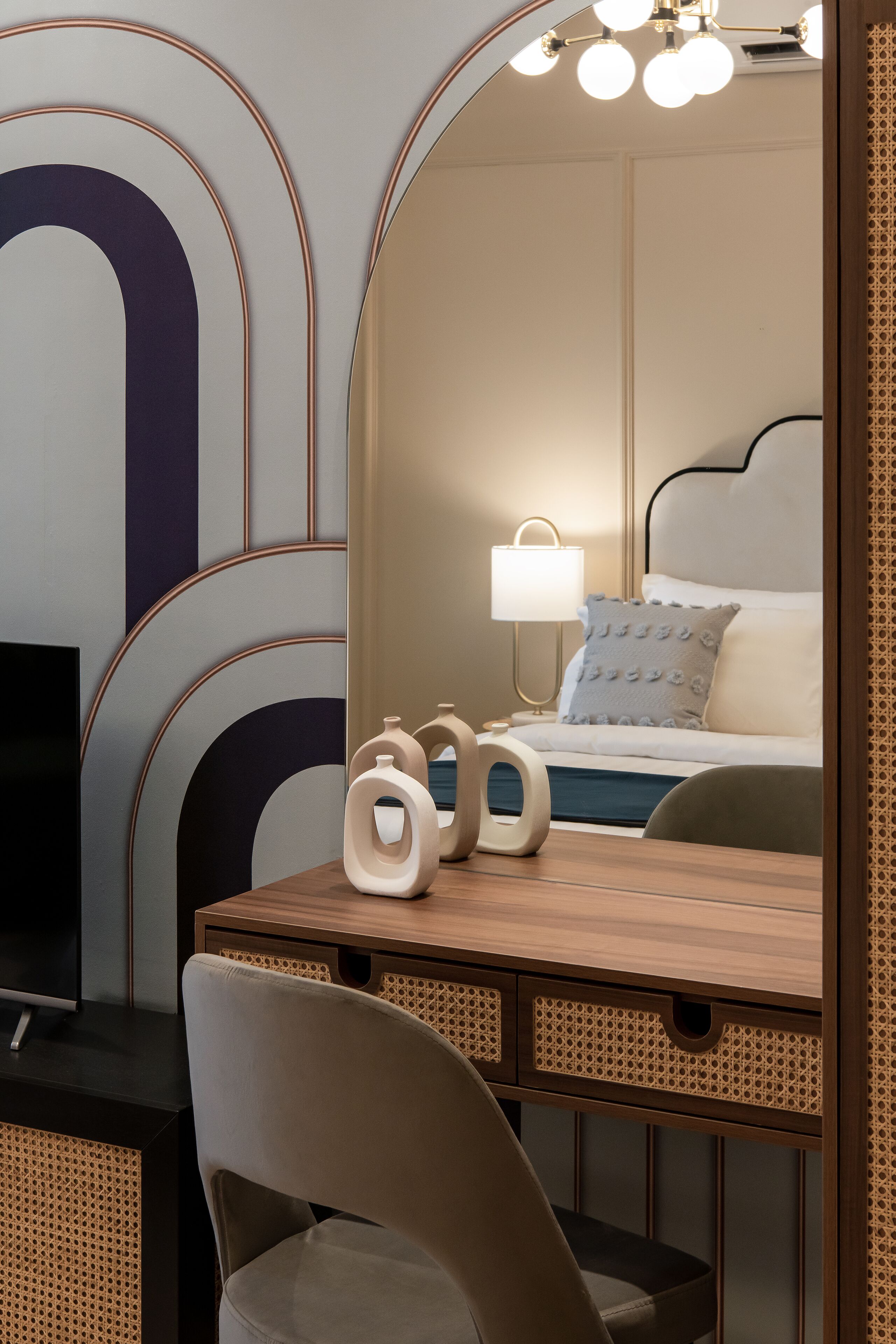 A modern hotel room with a wooden desk, a comfortable chair, a bed with a headboard, a bedside lamp, and a large mirror.