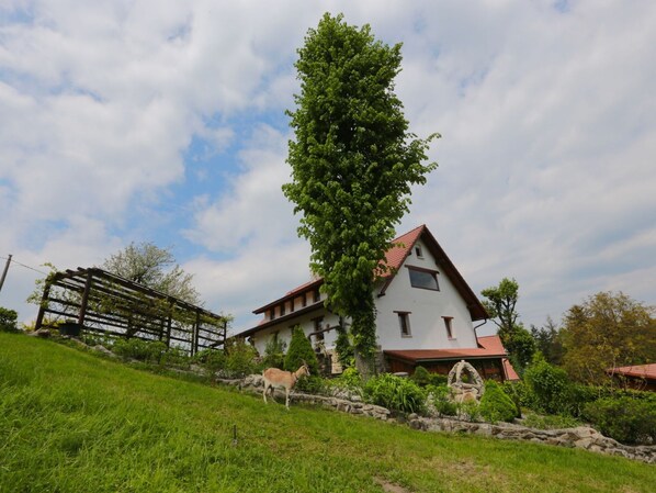 A two-story house with a red roof, surrounded by a lush green landscape and a large tree.