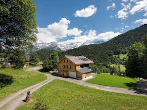 Une maison en bois avec un toit en pente, entourée d’un paysage verdoyant et avec des montagnes en arrière-plan.