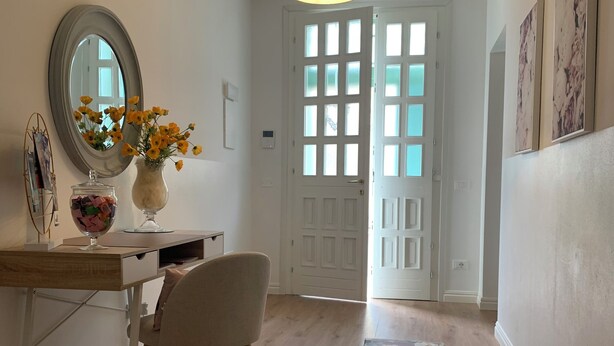 Un couloir doté d’un plancher en bois, d’un bureau avec une chaise, d’un vase de fleurs, d’un miroir et d’une porte dont les panneaux sont en verre dépoli.