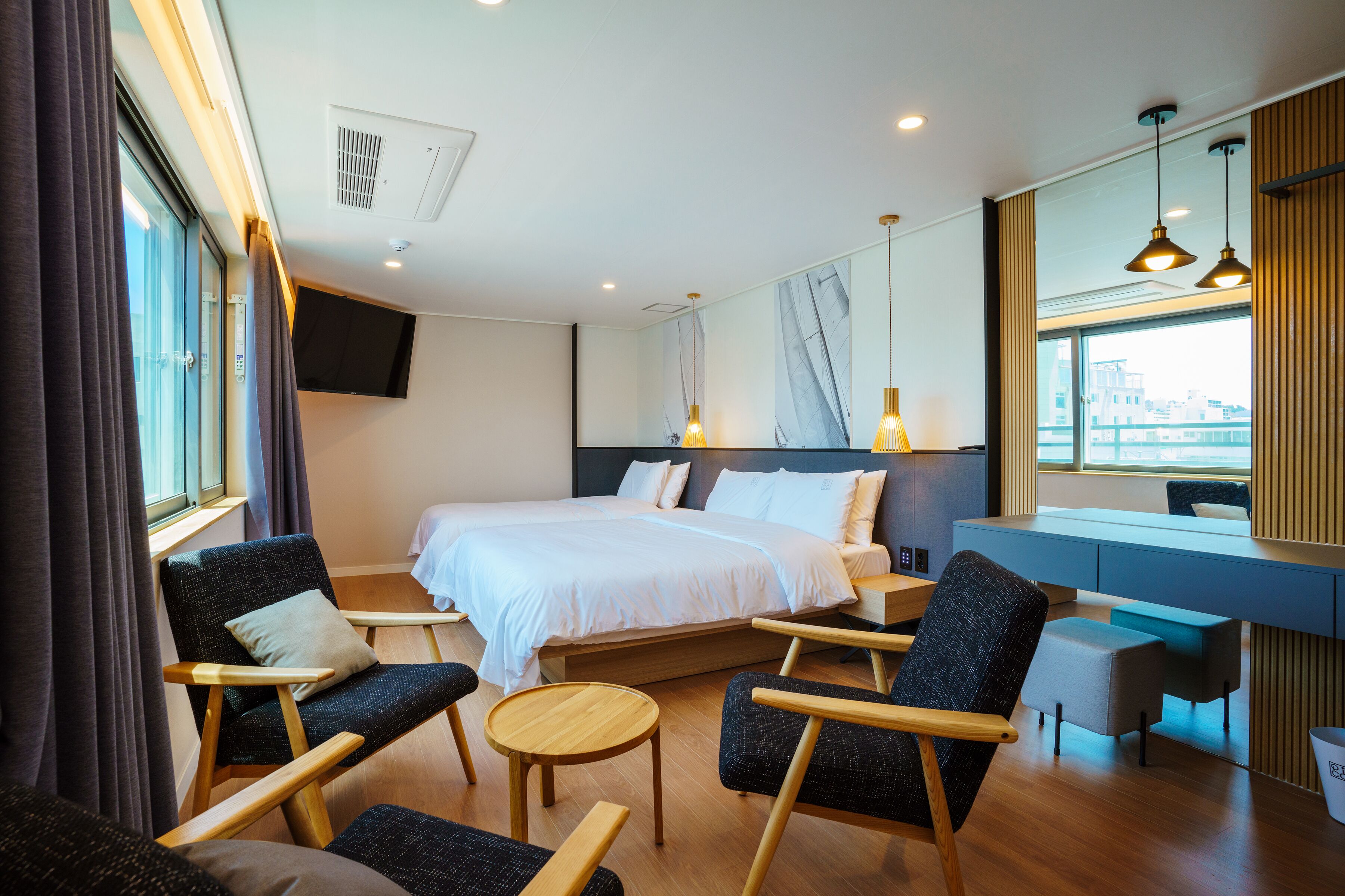 A modern hotel room with a large bed, two armchairs, a small round table, a desk, and a large window offering a city view.