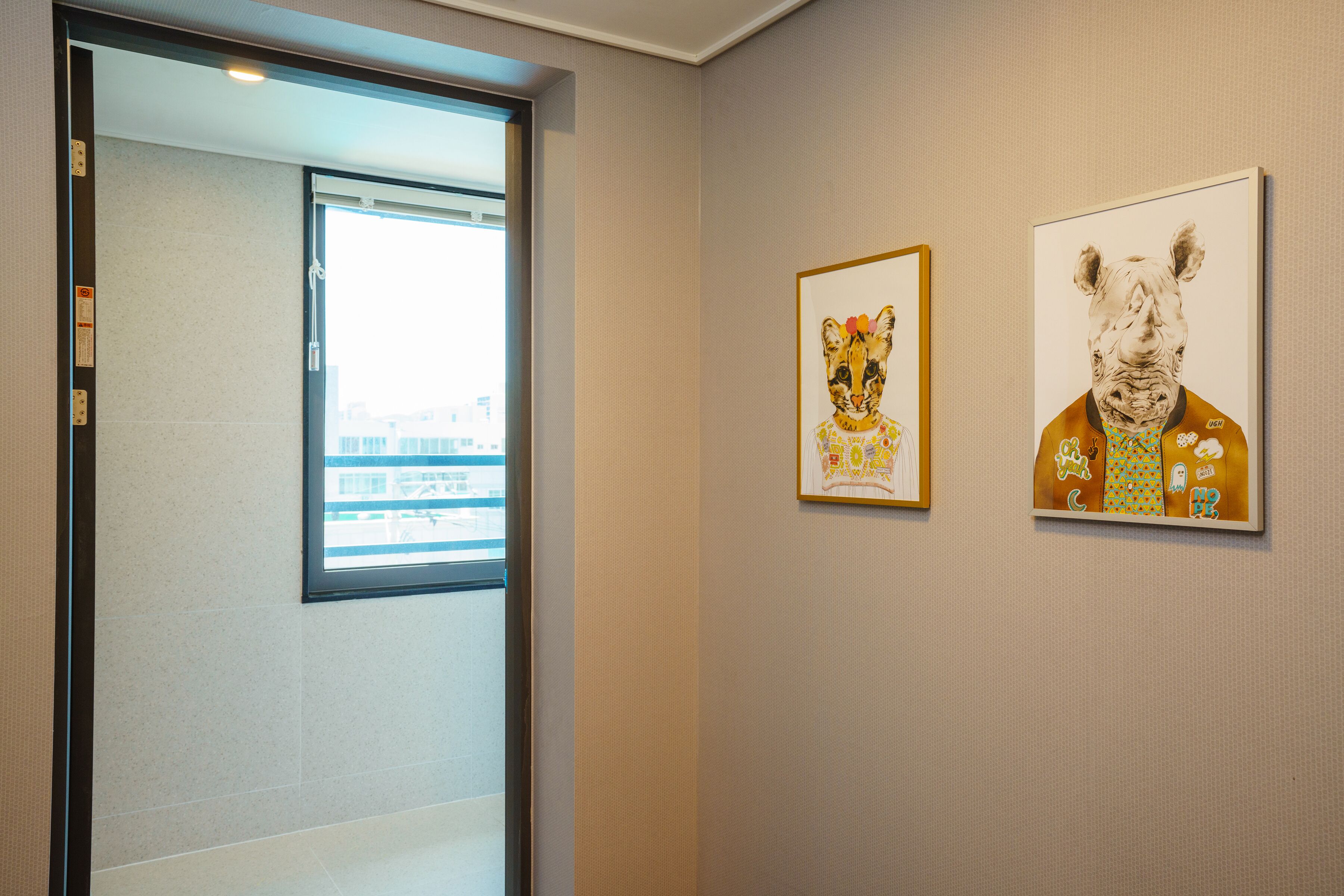 A hallway with two framed artworks on the wall, a window, and a door leading to another room.