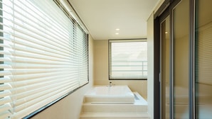 A modern bathroom with a bathtub, a window with blinds, and a glass door. - Hotel Pico (Tongyeong)