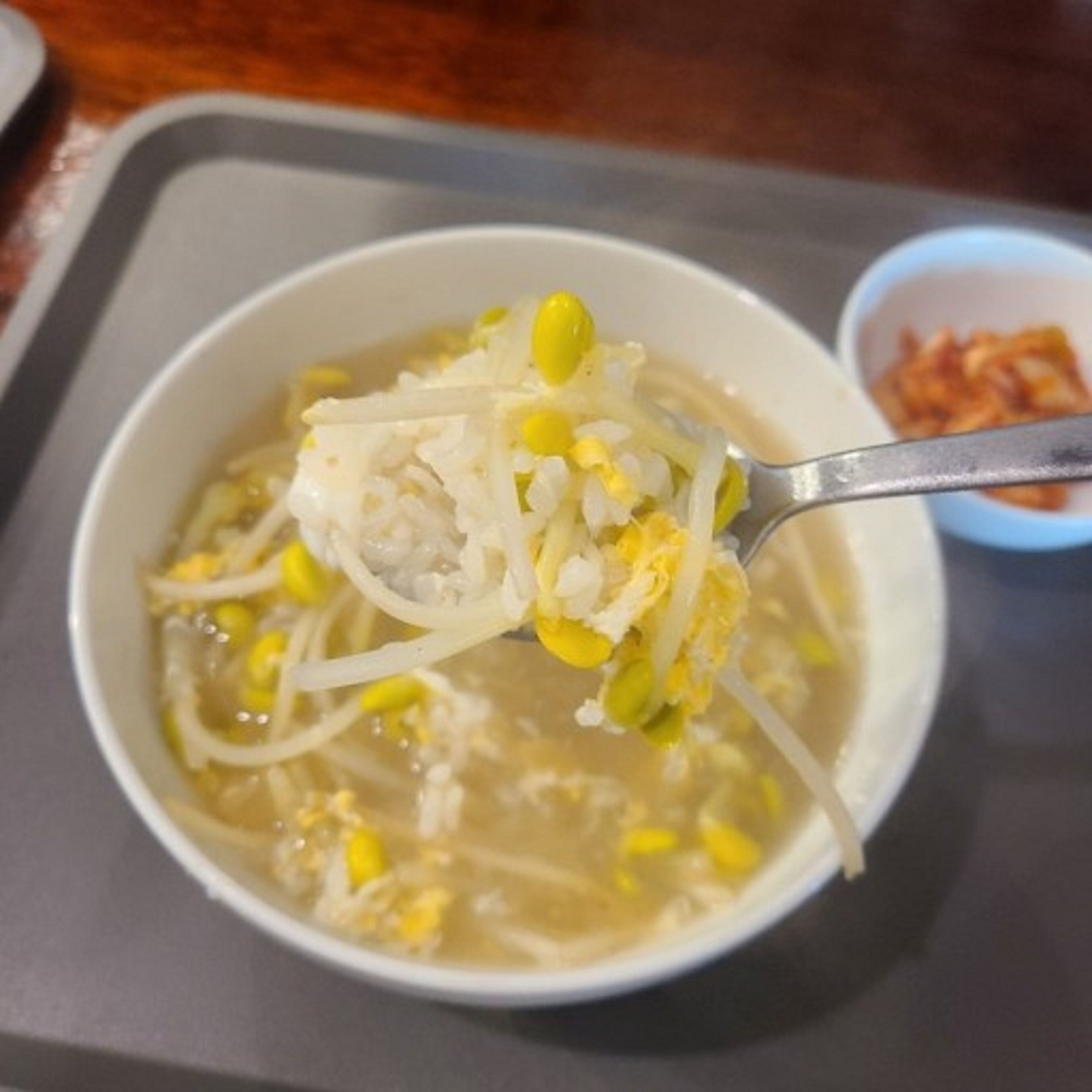 A bowl of soup with corn and shredded chicken, a spoon, and a small bowl of kimchi in the background.