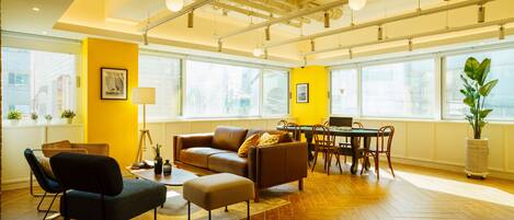 A modern living room with a herringbone wood floor, a brown leather sofa, a dining table with chairs, and large windows providing natural light.