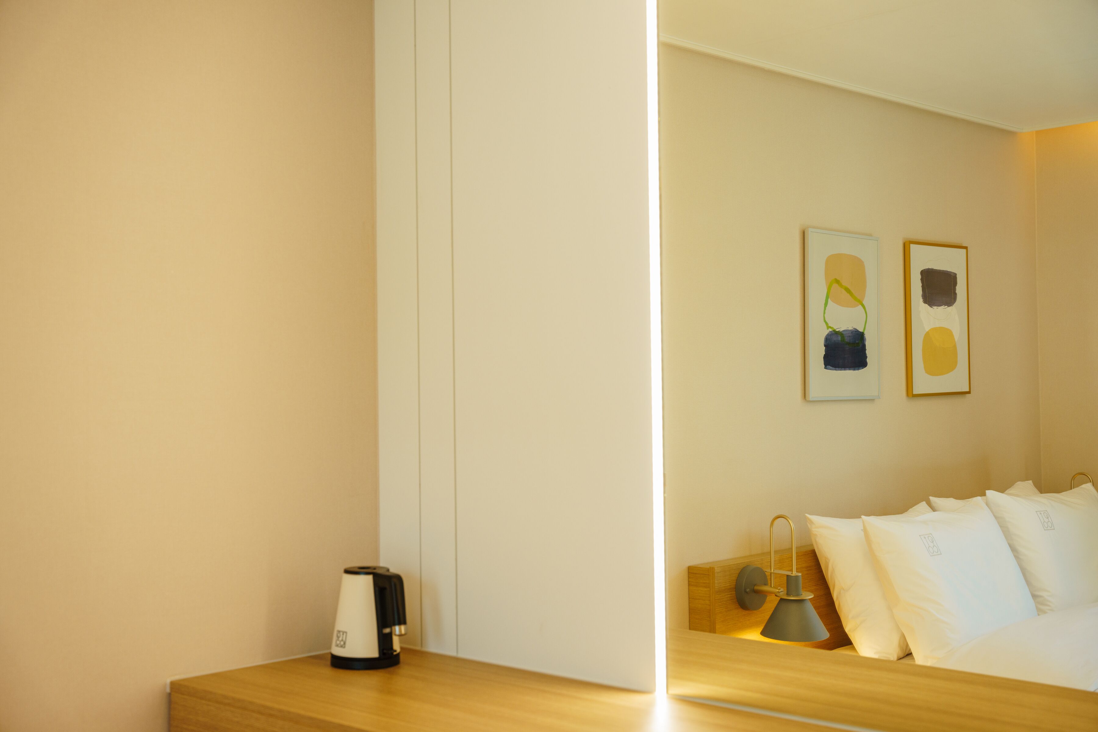 A hotel room with a wooden bedside table, a kettle, a lamp, and a bed with white pillows and a headboard.