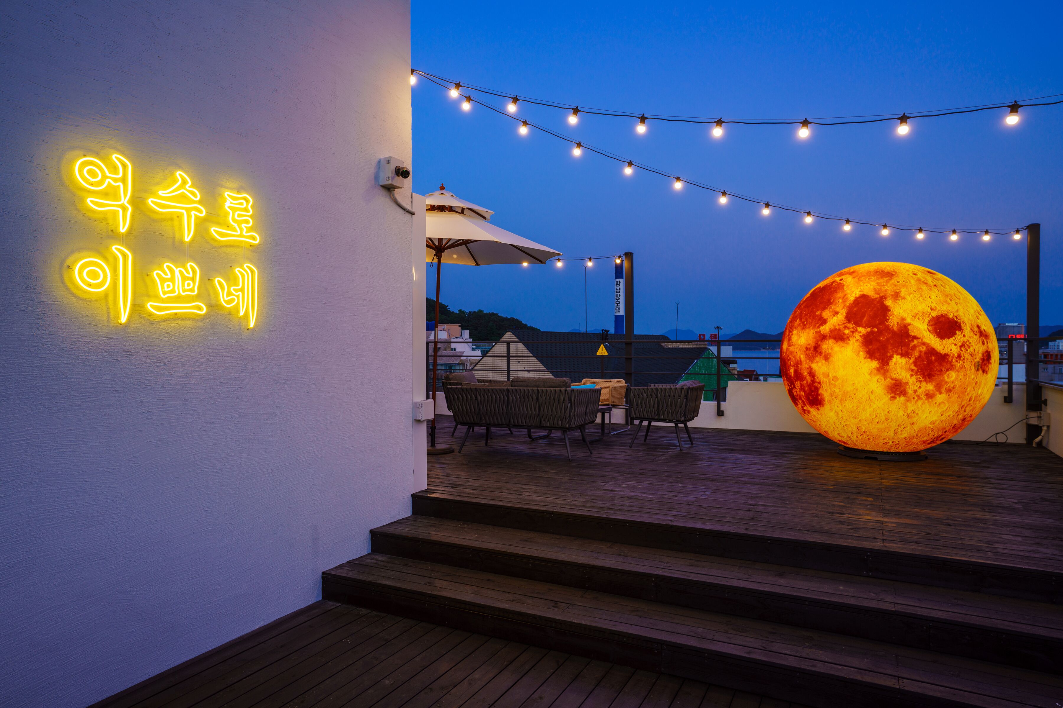A rooftop terrace with string lights, a large illuminated moon sculpture, and a neon sign in Korean.
