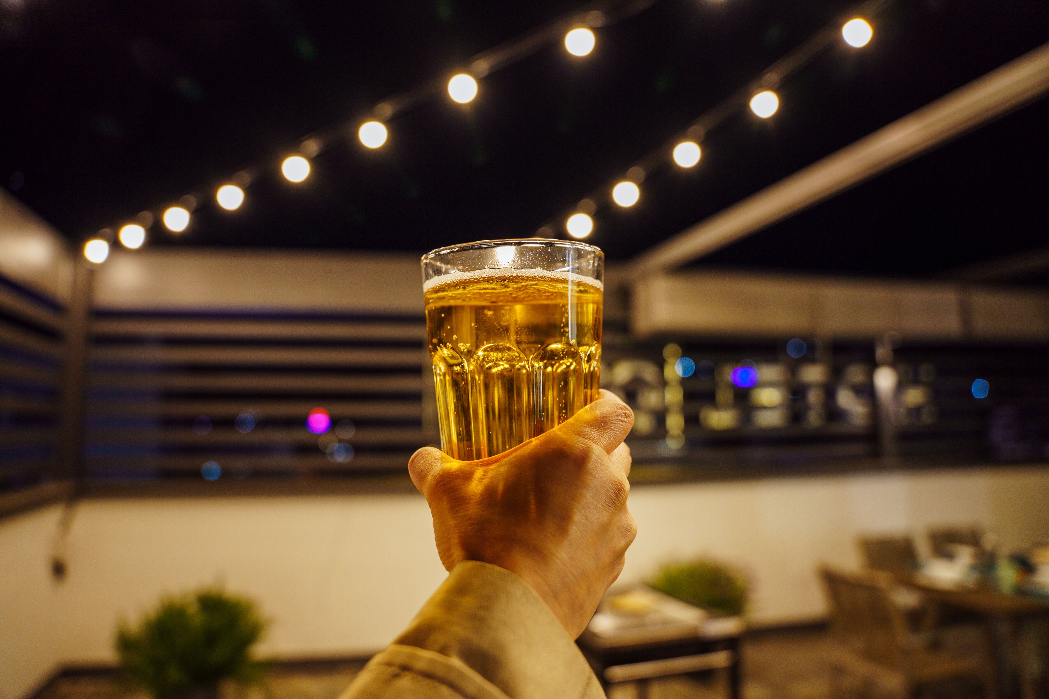 A hand holding a glass of beer in an outdoor setting with string lights in the background.