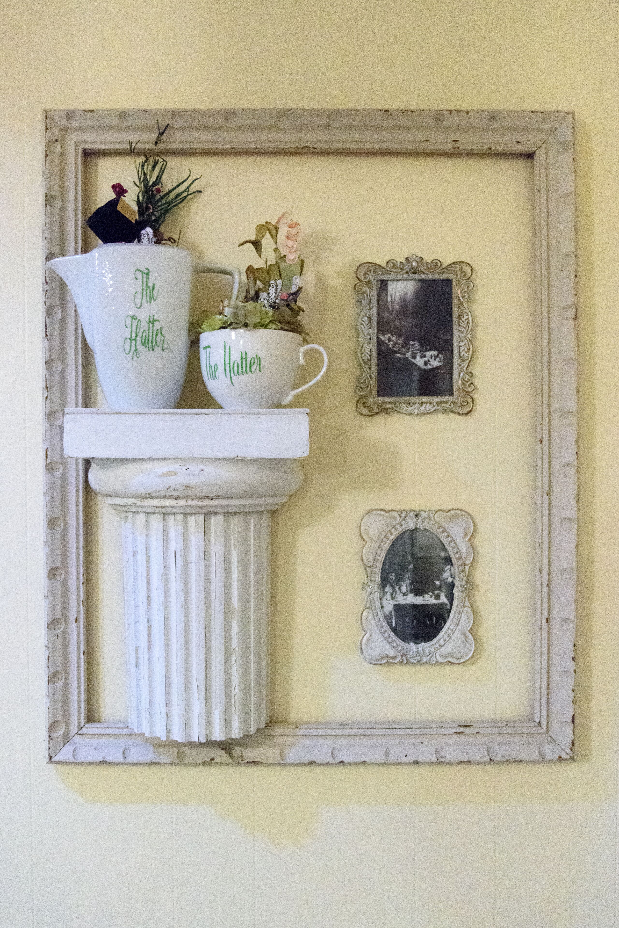 Ein an der Wand befestigtes Regal mit zwei weißen Krügen, auf denen 'The Hatter' steht, und einem kleinen gerahmten Bild.