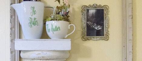 Ein an der Wand befestigtes Regal mit zwei weißen Krügen, auf denen 'The Hatter' steht, und einem kleinen gerahmten Bild.