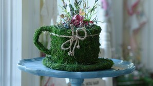 Ein mit Moos bewachsener Pflanztopf mit Blumenarrangements auf einem blauen Sockeltisch.