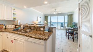 A kitchen with granite countertops, white cabinetry, and a dishwasher. There is a dining area with a table and chairs, and a view of the ocean through large windows.
