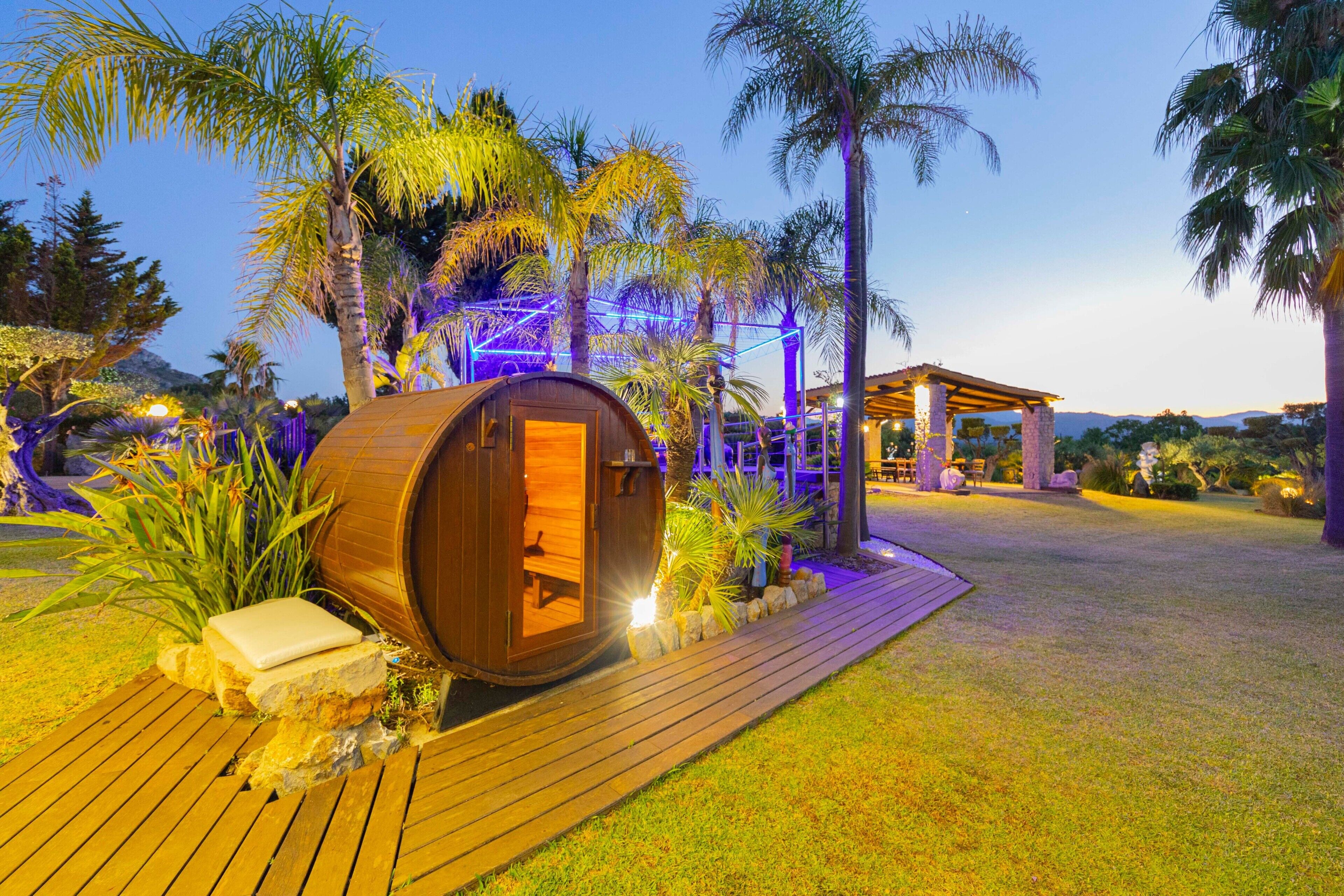 Une structure en forme de tonneau en bois, avec un banc à l’intérieur, entourée de plantes tropicales et dotée d’une terrasse en bois.