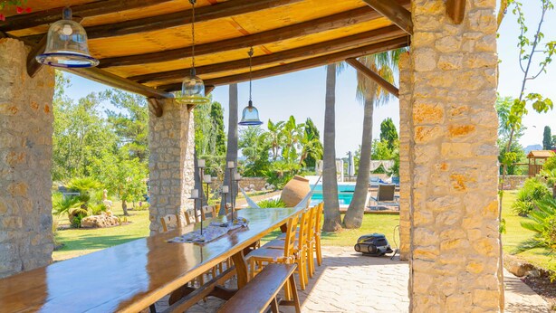 An outdoor dining area with a long wooden table and benches, stone pillars, hanging light fixtures, and a view of a swimming pool and greenery. - Country House 'Els Oscols 53' with Sea Views, Wi-Fi and Air Conditioning (Alcudia)